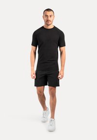 Camiseta negra de manga corta combinada con pantalones cortos negros, ambos hechos de una tela suave. Lleva puestas unas zapatillas deportivas blancas mientras está de pie sobre un fondo neutro.