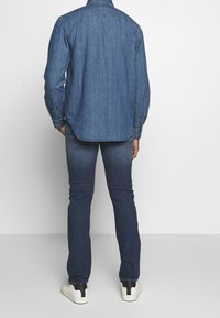 Camicia di denim blu medio, maniche lunghe con polsini a bottoni, taglio dritto, yoke posteriore, abbinata a jeans slim-fit blu scuro e sneakers bianche.