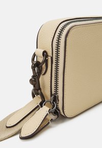 Beige läderplånbok med en texturerad yta, kontrasterande mörk dragkedja och metalldetaljer. Har två dragkedjepuller och en kompakt rektangulär form.