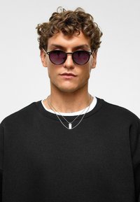 Zwarte sweatshirt gedragen over een witte T-shirt, met een ronde halslijn. Het model heeft krullend haar en draagt ronde, donkere zonnebril.