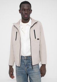 Veste zippée gris clair avec deux poches avant à fermetures éclair noires, une capuche et une texture lisse, associée à un jean en denim bleu.