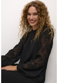 Black blouse with sheer, embroidered long sleeves. The neckline features a small button detail and gathers at the shoulders for texture.