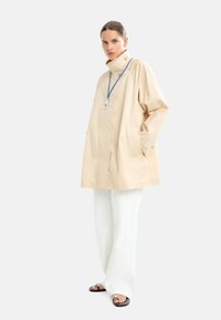 Beżowa, lekka kurtka z wysokim kołnierzem, z przodu na guziki oraz dwiema bocznymi kieszeniami, w zestawieniu z białymi spodniami typu wide-leg i minimalistycznymi sandałami.