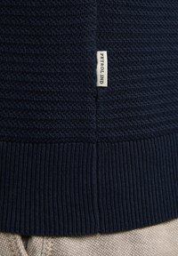 Pull en maille texturée bleu marine avec ourlet côtelé. Présente une petite étiquette blanche portant l'inscription "PETROLIND" près de la couture.