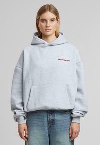 Felpa grigia in misto cotone, con tasca frontale, vestibilità oversize e testo rosso "PROIBITO" sul petto. Indossata con jeans azzurri chiari.