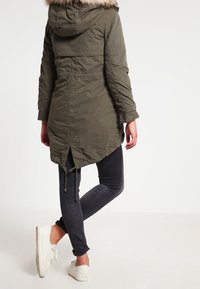 Olivově zelená parka s kapucí s umělým kožíškem, dlouhými rukávy, staženým pasem na šňůrku a zakřiveným lemem, doplněná černými džíny a bílými teniskami.