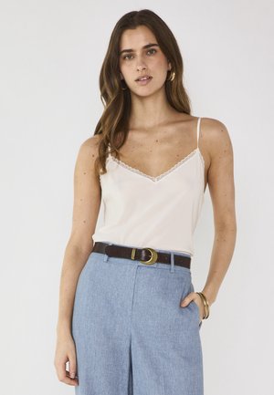 Donna con capelli lunghi castani che indossa una canottiera bianca con pizzo, pantaloni blu a vita alta, cintura marrone, orecchini a cerchio dorati e braccialetti.