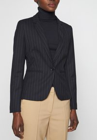 Kobieta ubrana w czarny golf i czarną marynarkę w prążki, z be�żowymi eleganckimi spodniami, stojąca na tle jasnej, jednolitej ściany.