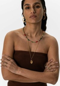 Gouden en kleurrijke kralenkettingen met een handpendant, gedragen met gouden oorringen en meerdere gouden ringen, tegen een neutrale achtergrond.