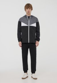 Giacca a vento nera e grigia con zip frontale, cappuccio e dettagli geometrici bianchi. Abbinata a pantaloni neri e sneakers bianche.