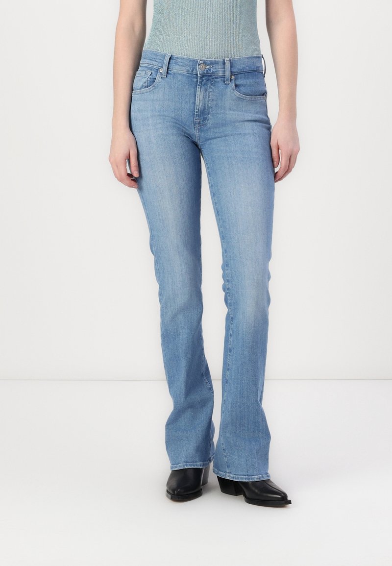 Personne portant un jean bootcut bleu clair, des bottes noires à bout pointu et un haut sans manches bleu clair, debout devant un fond blanc uni.