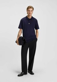 Navy polo shirt with short sleeves and a small logo, paired with black tailored pants and black shoes, holding a folded dark jacket.