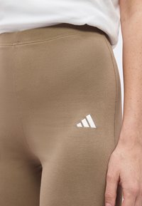 Gros plan sur un legging beige moulant avec le logo blanc Adidas sur la partie supérieure de la cuisse et la main d'une personne posée à côté.