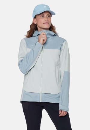 Kobieta w jasno niebiesko-białej kurtce zapinanej na zamek i pasującej czapce, poprawiająca kołnierz kurtki, stojąca na jednolitym tle.