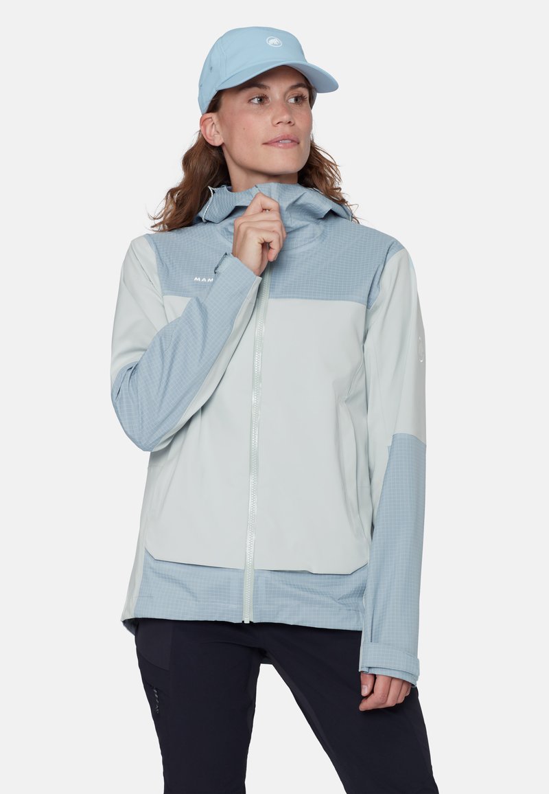 Frau trägt eine hellblaue und weiße Jacke mit Reißverschluss und passende Mütze, richtet den Kragen der Jacke, steht vor einem einfarbigen Hintergrund.