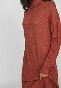 Pull à col roulé de couleur rouille, fabriqué en tissu tricoté texturé ; présente une coupe décontractée, des poignets côtelés et un design légèrement surdimensionné.