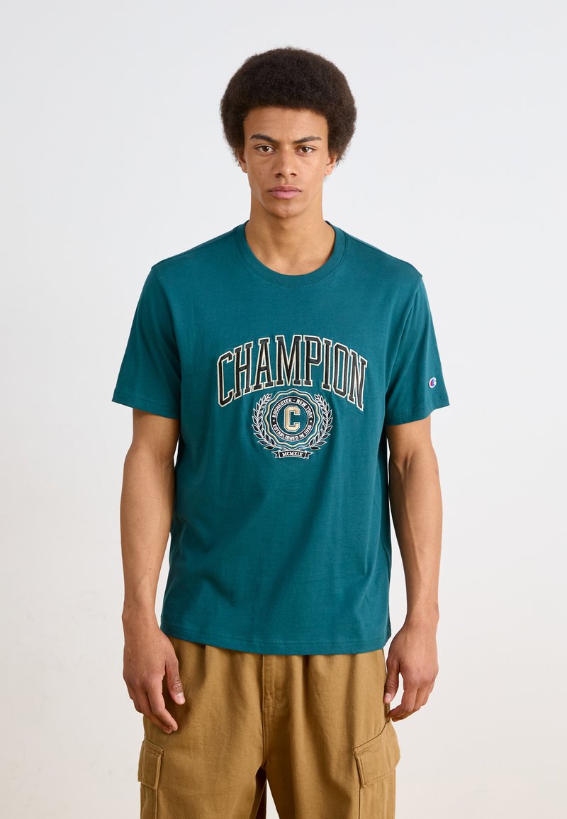 T-shirt de algodão turquesa com o logótipo "CHAMPION" em branco e preto, mangas curtas e decote redondo. Combinado com calções de carga bege.