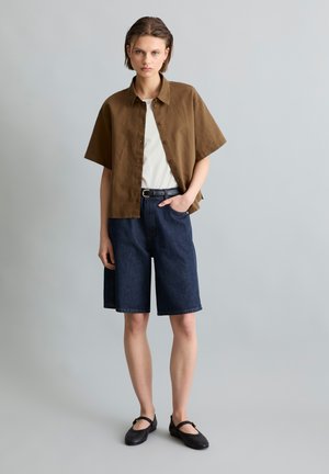 Junge Frau trägt braunes kurzärmeliges Shirt über weißem Top, dunkelblaue knielange Jeansshorts, schwarze Ballerinas, steht vor schlichtem grauem Hintergrund.