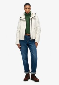 Biała kurtka z kapturem i czarnymi zamkami, zielony sweter z dzianiny, niebieskie dżinsy oraz brązowe sznurowane buty. Model stoi prosto, z rękami opuszczonymi wzdłuż ciała.