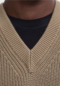 Dark-skinned person wearing a beige chunky knit V-neck sweater over a black crew neck shirt, cropped at the upper chest.