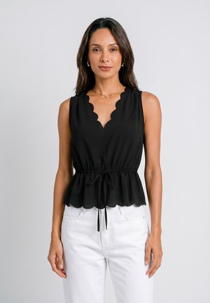 Vrouw met lang donker haar, die een mouwloze zwarte top met geschulpte rand en een taillekoord draagt, en witte broek, staand tegen een effen lichte achtergrond.