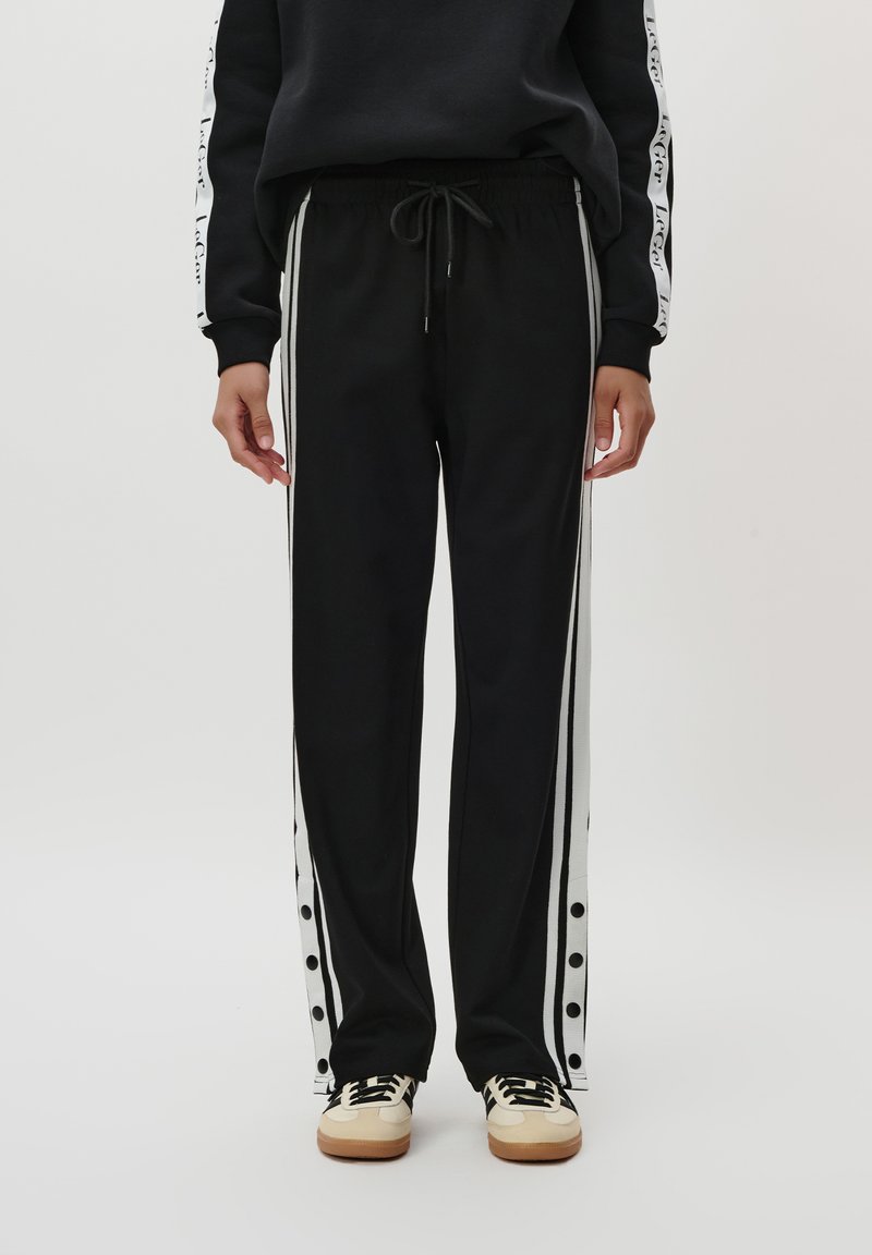Sorte joggebukser med hvite side-striper, med trykknapper langs sømmen. Laget av mykt stoff med elastisk midje og snøring.