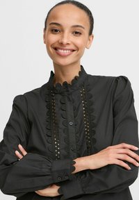 Black blouse with a high collar, scalloped trim, and decorative cut-out detailing. Features button fastenings and puffed sleeves.