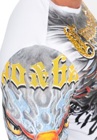 Weißes Kurzarmhemd mit einem auffälligen grafischen Design in Grautönen, Blau und Rot; gelber Schriftzug; sowie glitzernden Akzenten an der Schulter.