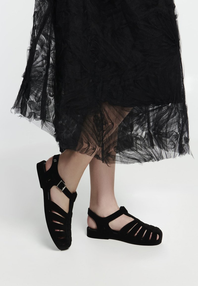 Sandales en suède noir avec design à bout ouvert, dotées de brides et d'une boucle à la cheville, associées à une jupe noire transparente et texturée.