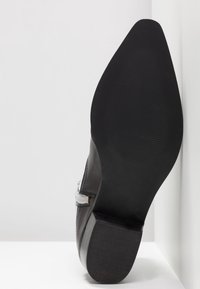 Semelle d'une chaussure habillée en cuir noir avec une boucle, inclinée contre un mur d'angle blanc et un sol.