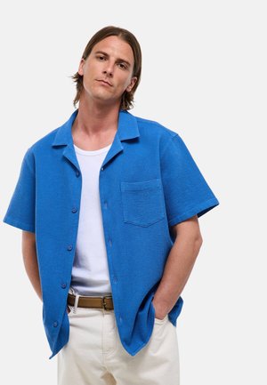 Homme aux cheveux mi-longs portant une chemise bleue à manches courtes boutonnée sur un t-shirt blanc et un pantalon beige, les mains dans les poches.