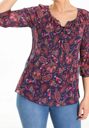 Woman wearing a navy blue floral blouse with red and pink patterns and light blue jeans, arm raised, cropped at neck and hips.