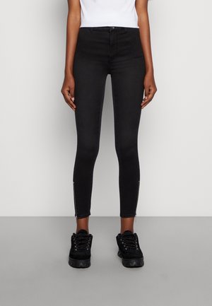 Femme portant un jean skinny noir avec des zips aux chevilles, associé à des chaussures à plateforme noires à lacets, se tenant sur un sol gris avec un fond de mur blanc.