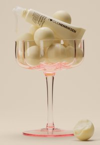 Una coppa in vetro trasparente con gambo rosa contiene sfere beige e un tubo di trattamento per labbra Ole Henriksen, che gocciola lievemente, su uno sfondo beige.