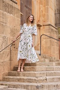 Femme en robe midi fleurie tenant la rampe d'escalier tout en se tenant sur des marches en pierre à l'extérieur d'un bâtiment historique aux murs texturés et à la porte en bois.