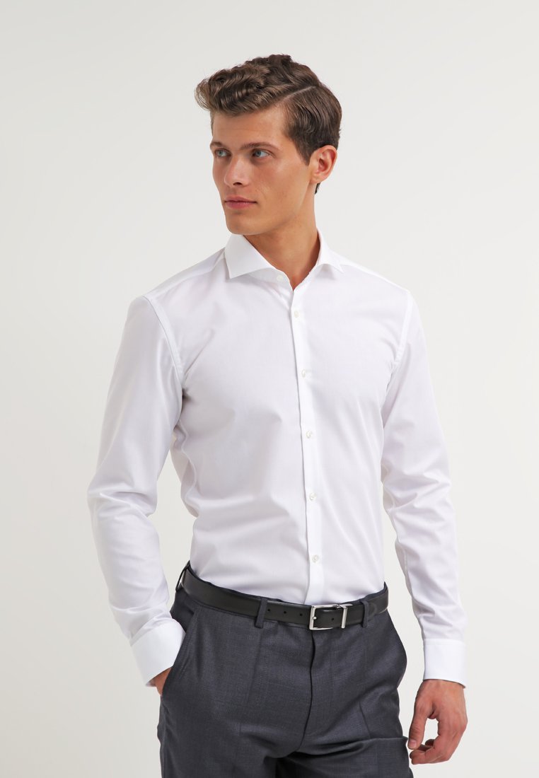 White long-sleeve shirt with a pointed collar, buttoned front, and cuff detail, paired with dark tailored trousers and a black belt.