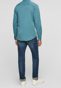 Chemise turquoise à manches longues avec poignets à boutons, associée à un jean bleu foncé. Baskets blanches visibles à la cheville, texture lisse, design décontracté.