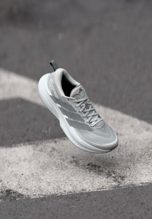 Lichtgrijze sportieve sneaker met witte zool en veters, zwevend boven een asfaltweg met witte pijlaanwijzing.