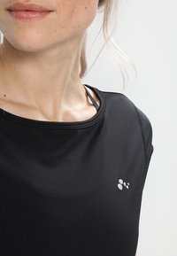 Camisa atlética negra con un acabado texturizado, que presenta un cuello redondo y un pequeño logo en plata reflectante en el lado inferior.