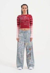 Červený dlouhý rukáv crop top s grafickým potiskem přechází do světle růžových rukávů; široké světle modré džíny s propracovanou výšivkou květin a vzorů.