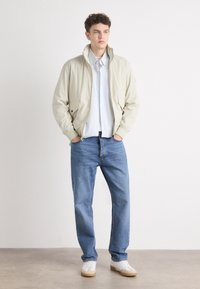 Lyse beige bomberjakke med glidelås foran, ribbestrikkede mansjetter og sidelommer, båret over en stripet skjorte og blå jeans med hvite joggesko.