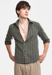 Maglietta a righe verde oliva in tessuto leggero, con scollatura a V profonda, maniche arrotolate e bottoni marroni. Indossata con pantaloni nei toni neutri.
