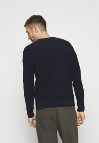 Pulover tricotat bleumarin cu mâneci lungi, guler rotund și tiv ribat. Purtați peste pantaloni verzi măslinii, evidențiind designul texturat al spatelui.