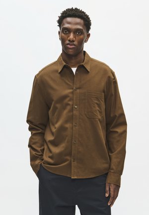 Homme portant une chemise marron boutonnée avec une poche poitrine et un pantalon noir, debout devant un fond blanc.