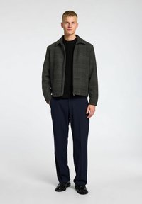 Veste à carreaux gris avec col, tissu texturé, intérieur en tricot noir, portée avec un pantalon bleu marine foncé et des chaussures noires.