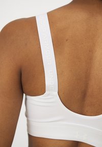 Balta sportinė liemenėlė su plačiomis petnešomis, ant kurių užrašyta "UNDER ARMOUR". Lygus audinys su apvalia nugaros konstrukcija, minimaliais siūlėmis ir logotipu.