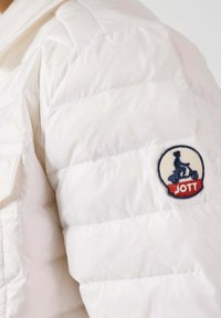 Giacca bianca trapuntata con una piccola toppa circolare con il logo "JOTT" e un'illustrazione di una persona su un monopattino. Materiale leggero e testurizzato.