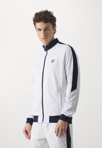 Weiße Trainingsjacke mit marineblauen Akzenten, hohem Kragen, Reißverschluss und Logo auf der Brust. Glattes Material mit gerippten Bündchen und Saum.