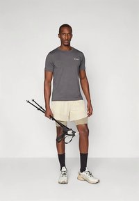 Αθλητικός άνδρας με γκρίζο πουκάμισο και μπεζ σορτς κρατάει μαύρες πεζοπορικές ράβδους, φοράει μαύρες κάλτσες και μπεζ μποτάκια πεζοπορίας, στέκεται σε γκρίζο δάπεδο.