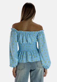 Blusa blu off-the-shoulder con vita arricciata, maniche lunghe, motivi floreali gialli e morbida texture di cotone. Abbinata a jeans chiari.